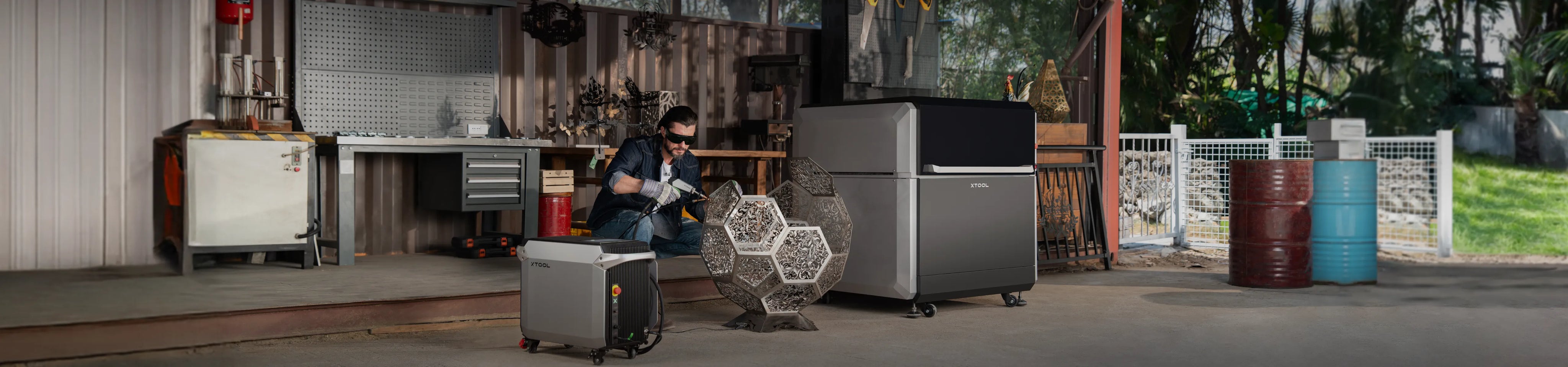 A man in a workshop wearing a protective face mask and suit works beside a large stainless steel refrigerator and a smaller gray appliance. The scene is set in a real industrial workshop environment with tools, barrels, and outdoor greenery visible in the background.