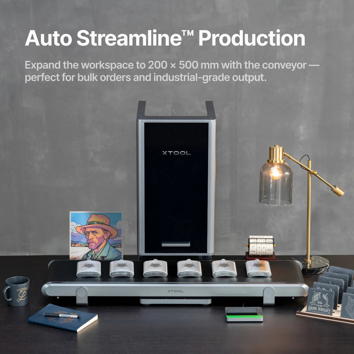 The front of the xTool F2 Ultra with the conveyor attached showing finished products including whiskey flasks, coasters, a mug, portrait and notebook.